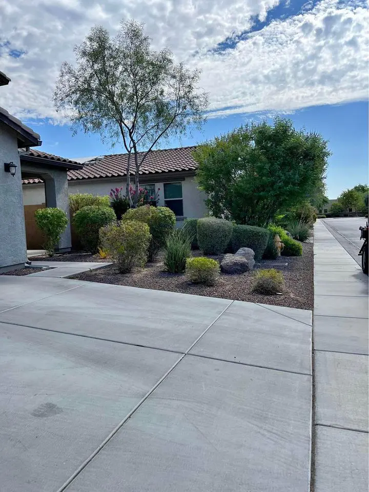 A house with a tiled roof and light-colored walls is surrounded by a beautifully designed garden featuring bushes, trees, and rocks. Thanks to expert landscape maintenance, the Phoenix landscaping shines under the partly cloudy blue sky along the long driveway extending from the front.